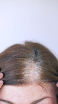 Vertical video. Close view of gray hair at the scalp as woman reveals gray hairs along roots. Clip shows natural color transition, authentic texture and aging signs. Brunette with gray roots.