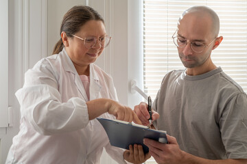 male patient, bald man hospital, medical consultation patient and doctor, documenting, patient reviewing, signing medical forms, routine checkup, writing notes on clipboard, diagnosis and examination
