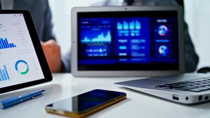 A man in a grey suit is displaying charts on various devices like tablet and laptop. A smartphone is in the foreground - Powered by Adobe