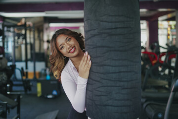Young woman smiling and expressing confidence while standing next to a boxing punching bag during a workout session