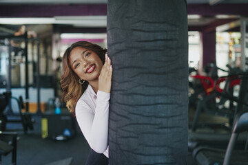 Fototapeta premium Young woman smiling peeking from behind punching bag in gym