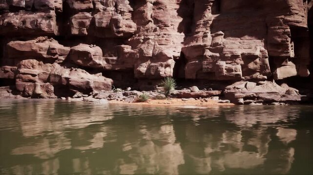 The Colorado River flows gently beside towering rocky cliffs, reflecting the morning light. A small patch of land with greenery emerges along the riverbank, creating a serene atmosphere.