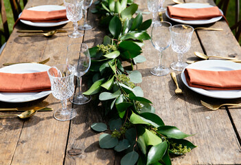 Rustic wooden table setting with greenery garland and terracotta napkins. Outdoor wedding reception decoration with gold cutlery and crystal glasses. Boho chic dining arrangement