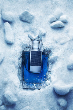 A bottle of fragrance is placed inside ice and snow. The setting shows a mix of icy surfaces and natural stones. The scene captures a moment of cold beauty.
