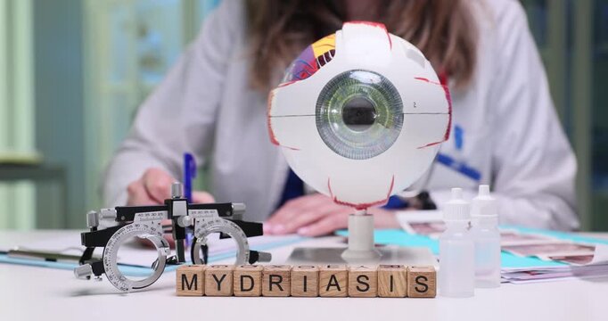Woman ophthalmologist carefully writes prescription for vision medications. Miniature wooden cubes arranged neatly on table form word Mydriasis