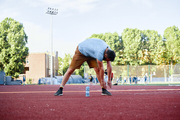 A man performs a stretching exercise on a sport's track in a vibrant outdoor sports facility surrounded by people and tall trees showcasing physical activity and healthy lifestyle.