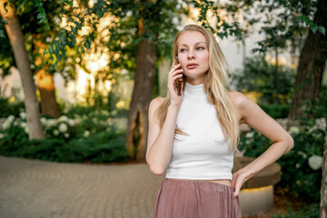 A beautiful blonde woman is talking on the phone in a city park. Communication, business relations.