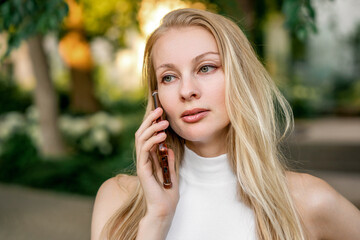 A beautiful blonde woman is talking on the phone in a city park. Communication, business relations.