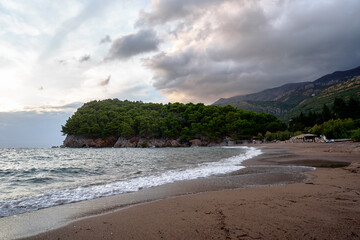 A picturesque beach with gentle waves, a forested island, and dramatic clouds creating a peaceful ambiance of nature's beauty meeting the sea.