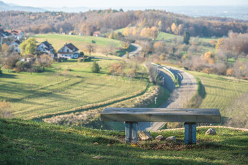 Ruhebank am Wanderweg Gustav-Str&ouml;hmfeld-Weg und Eduard M&ouml;rike Weg mit Aussicht auf herbstliche Landschaft mit geschwungener Stra&szlig;e und Ortschaft Kappish&auml;usern  am Jusiberg in Kohlberg im Streiflicht.