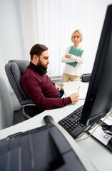 A bearded man sits at a white desk, writing on documents, as a blonde woman stands nearby holding green folders, observing in a bright modern office. Computer, printer and papers surround them.