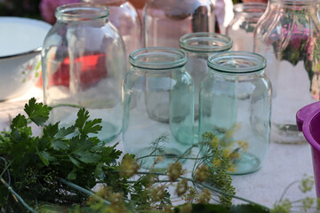 Close-up of empty glass jars on a table with fresh green herbs in the foreground. Bright natural light creates reflections on the glass, adding a clean and rustic feel. home canning