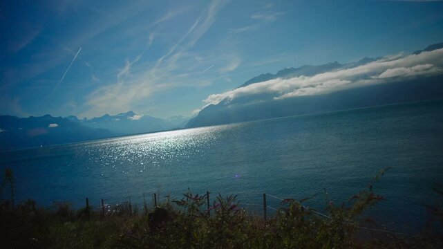 Bright sunlit lake with shimmering water and distant mountains seen from moving train window creating peaceful scenic travel moment with clear sky clouds and natural beauty