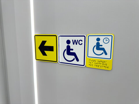 Set of signs for people with disabilities on wall. Arrow points to accessible restroom and to waiting area with braille text. Directional navigation for people with mobility issues