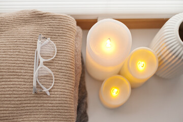 Pile of warm knitted sweaters, led battery candles and vase over white background with soft window light.