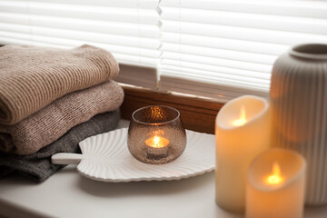 Pile of beige knitted sweaters, wooden tray, candles and vase over white background with soft window light