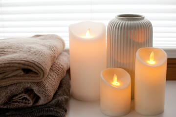 Pile of warm knitted sweaters, led battery candles and vase over white background with soft window light.