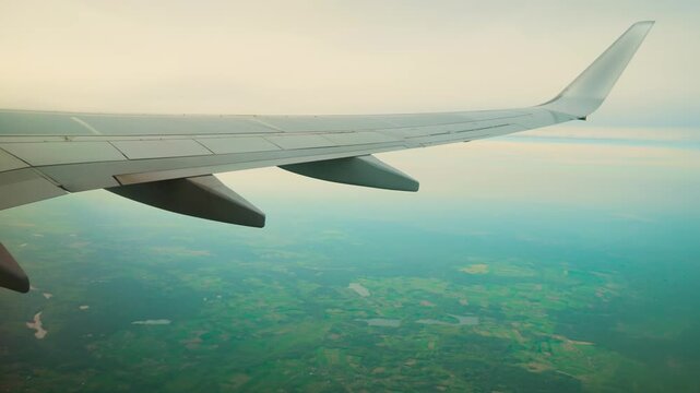 Airplane wing flying above land, flaps ailerons winglet over green fields. Aircraft wing gliding through sky, flaps spoilers tip above countryside. Jet wing cruising high, control surfaces winglet