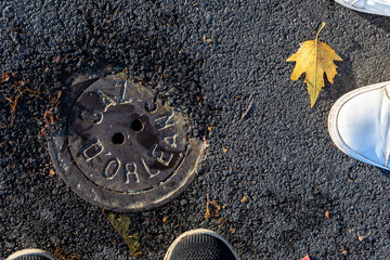 Round metal manhole cover with text Gaz d Orleans on asphalt street ground. Urban utility hatch detail in Orleans France with yellow autumn leaf and shoes on a sunny day from above
