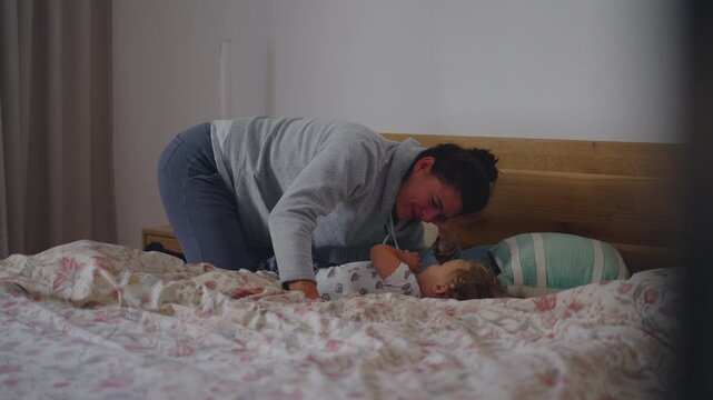 Mother leaning over toddler on bed during playful morning moment showing warmth, laughter, and energetic bonding in cozy home setting while beginning relaxed family start to day together