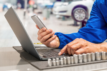 Close-up of hands of manager at the auto repair shop  in a blue uniform a car engine, engaging with his mobile phone in a garage environment, embodying satisfaction and modern technology at work