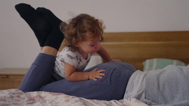 Playful toddler climbing onto mother&rsquo;s back on bed, balancing briefly before tumbling in warm family moment filled with joy, bonding, movement, spontaneity, and affectionate interaction