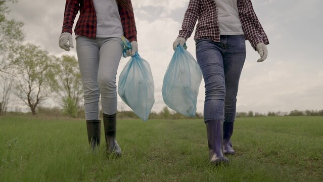 Girls walk on a green field in rubber boots and hold garbage bags in their hands, ecology of cleanliness, volunteer awareness, pollution, environment, happy family, plastic garbage, waste