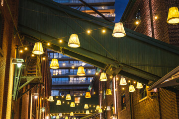 Fototapeta premium Hanging decorative lamps and warm string lights between brick buildings in Torontos Distillery District at night.