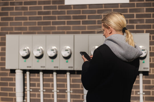 Middle-aged woman checking multiple outdoor electricity meters and entering readings on her smartphone beside a brick building
