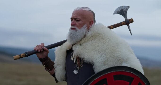 Viking warrior with axe standing on mountain cliff, wearing fur cloak and leather armor, overlooking dramatic landscape. Historical Nordic fighter, strength and adventure concept. Slow motion