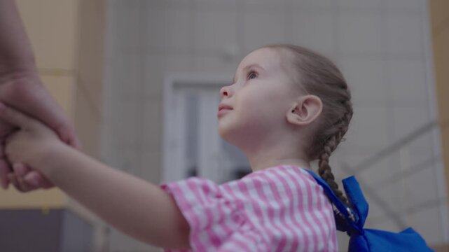 little girl with backpack holds her mother by the hand and goes to school, kid with mom go to school in the school yard, child with school bag hurries to first lesson, preparatory preschool education.