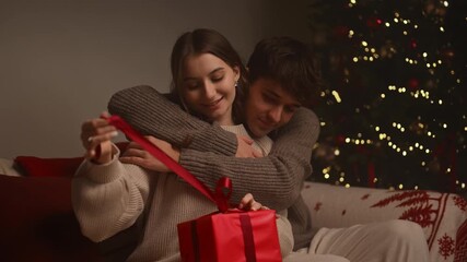 A warm holiday moment as a young couple sits embraced on a cozy sofa, the young woman happily unwrapping a red gift box while glowing Christmas lights create a festive, intimate atmosphere. - Powered by Adobe