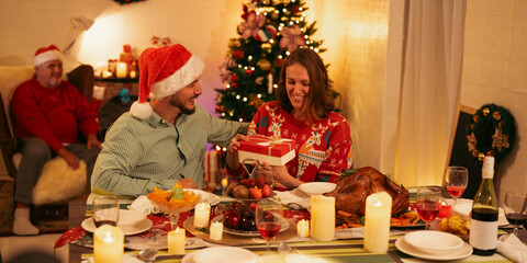 Romantic couple exchanging Christmas gift box at festive dinner table with Christmas tree and holiday decorations background, joyful celebration, love, happiness