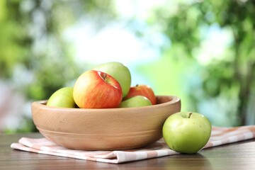 Fresh apples and bowl on wooden table against blurred green background, closeup