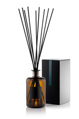 A brown glass bottle holds black reeds for scent release, standing beside a simple black and white package, placed on a clean white surface for enhancing indoor aroma.