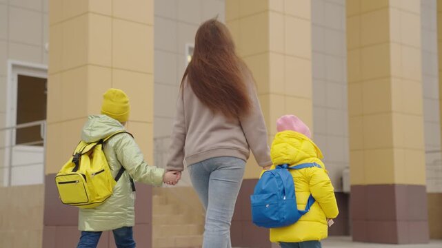 mother holds children with backpacks by hand and leads them to school, primary school students rush to class, mother and daughters girls go to first grade, school bag on her shoulders behind her back.