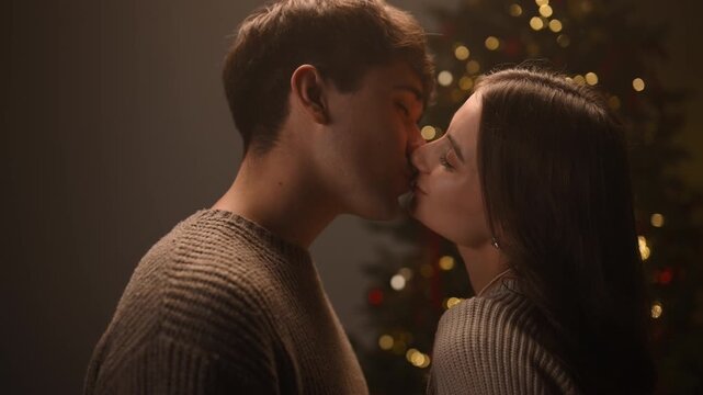 Romantic moment of a couple leaning in for a tender near-kiss, then kissing softly, surrounded by warm Christmas lights and a decorated tree creating a cozy festive atmosphere.