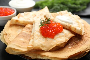 Delicious crepes with red caviar and sour cream on black table, closeup