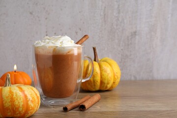 Delicious pumpkin latte with whipped cream in glass cup, cinnamon sticks, fresh vegetables and burning candle on wooden table against grey background, closeup. Space for text