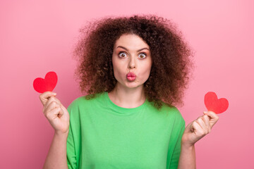 Young woman with curly hair in a green tshirt holds red hearts and gives a playful kiss against a pink background for love lifestyle advertising