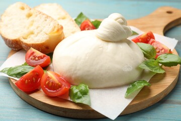 Delicious burrata cheese with tomatoes, bread and basil on light blue wooden table, closeup