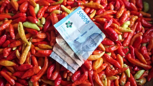 Close-up of a cash transaction using Indonesian rupiah at a traditional market, with hands exchanging money above a pile of fresh chili peppers. Perfect for themes of trade, economy, finance