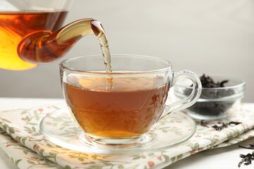 Pouring hot tea into cup from teapot at white table, closeup