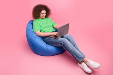 Young woman sitting on a blue bean bag using a laptop smiling in a bright green tshirt on pink background