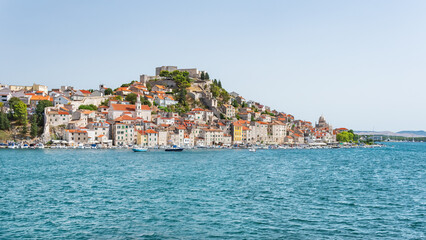 The coastal town of Sibenik in Croatia, nestled on the seafront with hilltop houses and St. Michael's Castle, is seen from a walking path on a sunny summer day.
