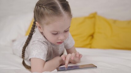 little child plays fun phone game lying on bed in room, a happy kid learns through smartphone application, children's education online through gadget sensor, girl daughter does homework with pleasure.