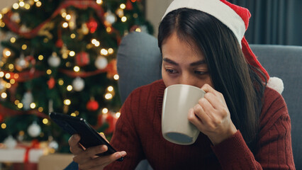 Positive Asia female wear red sweater new normal celebrate christmas smell aromatic coffee cup sit on wing chair check online greeting message from friend and family on phone in living room at house.
