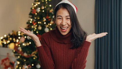 Young attractive Asia female blogger looking at camera video call say merry christmas and happy new year, Christmas tree decorated with ornament in living room at home. Holiday festival concept.