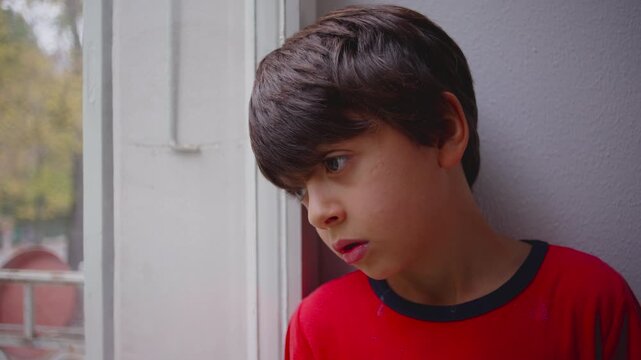 Boy standing quietly by window with thoughtful expression while gazing outside creating calm indoor moment of introspection curiosity and stillness framed by soft natural light