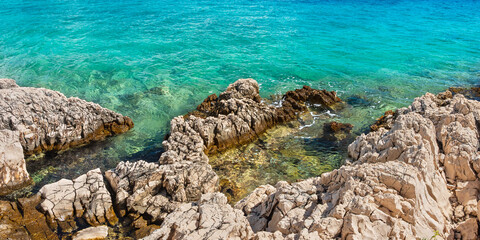 Natural shoreline with sharp rocks jutting out above the clear blue water, viewed from the shore on a sunny day.